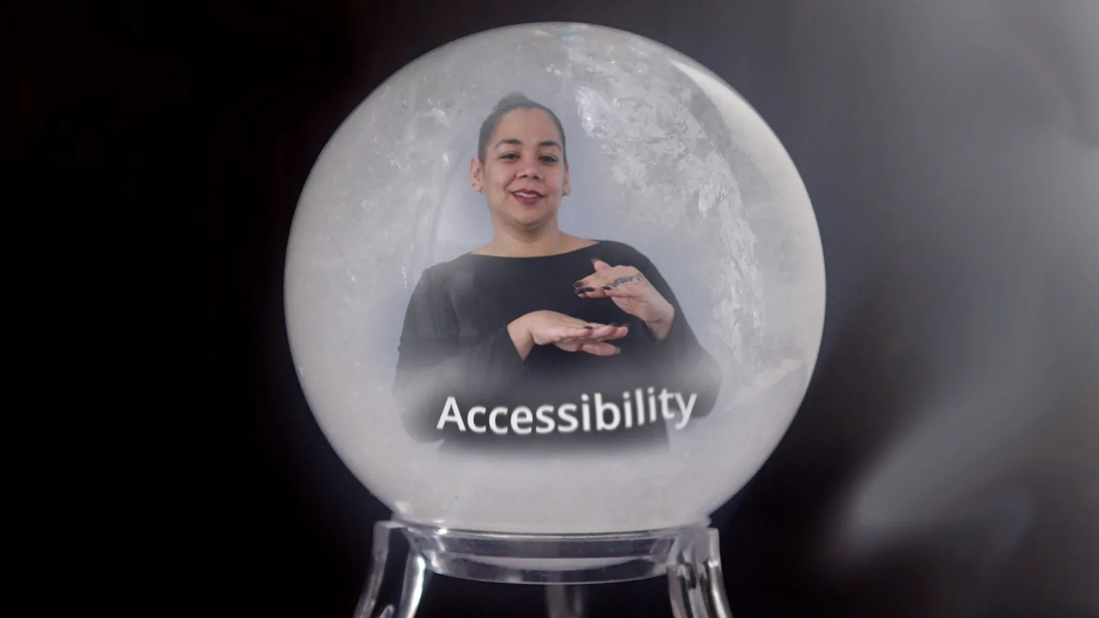 The image showcases a snow globe effect featuring a woman who appears to be signing, indicating the concept of "Accessibility." The woman is situated in the center, dressed in a dark outfit, with her left hand raised and her right hand positioned below it, creating a gesture that likely represents communication or signing. The background shows a softly blurred snowy texture, enhancing the wintery aesthetic of a snow globe. The word "Accessibility" is prominently displayed at the bottom of the globe in white text, making it a focal point. The overall color scheme combines dark tones with lighter shades creating a contrast that emphasizes the subject matter.