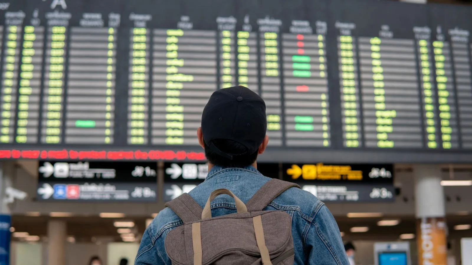 Person in a denim jacket and cap looking at a flight information board in an airport terminal.