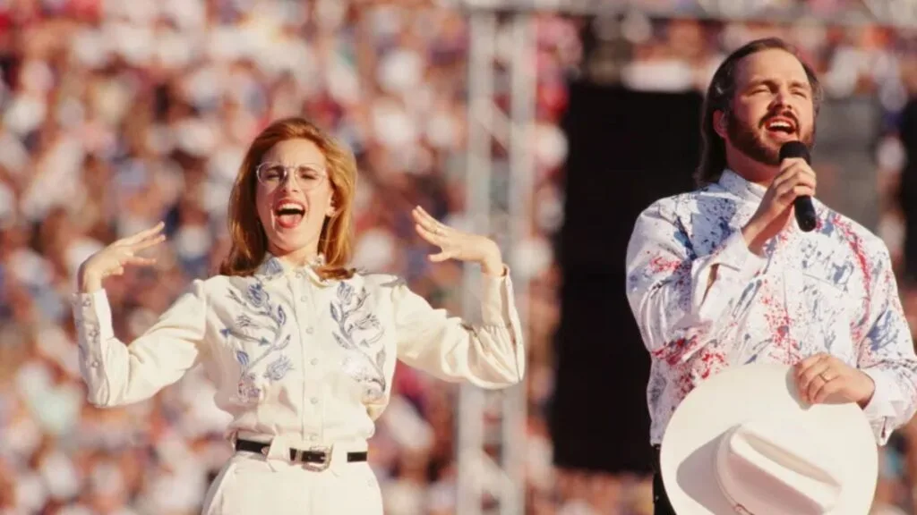 Marlee Matlin, is passionately signing the national anthem while wearing a white, Western-style outfit with blue embroidery. Beside her, country singer Garth Brooks is singing into a microphone, wearing a white cowboy hat and a white shirt with red and blue paint splatters. The background shows a blurred stadium crowd.