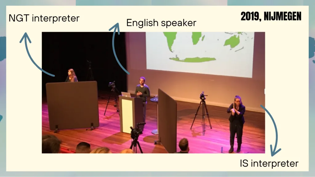 On a conference stage, an English speaker stands at a podium and addresses the audience through a microphone. The layout includes an NGT interpreter positioned on the left and an IS interpreter on the right. Strategically placed screens serve to section off each of the three professionals currently on stage.