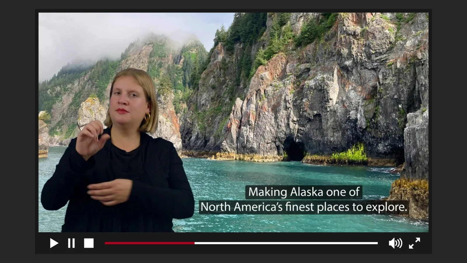 A travel tour video showing the rugged, steep cliffs and vibrant turquoise waters of Alaska's coastline. This video includes captions and a sign language interpreter, dressed in black.